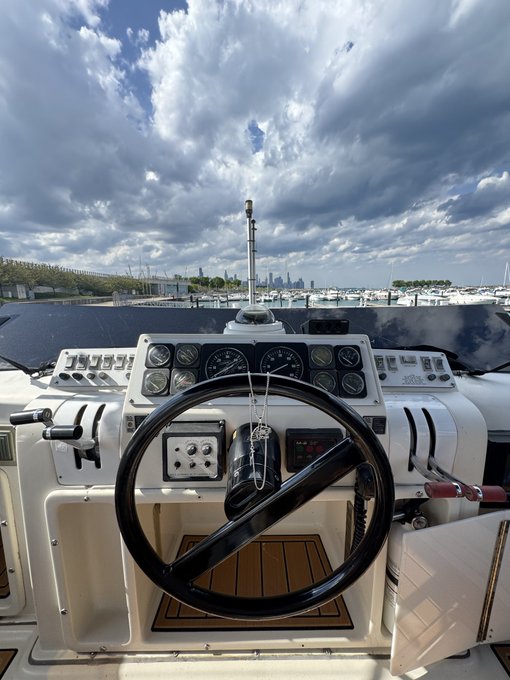The helm and dashboard of a boat are visible, featuring a black steering wheel and multiple gauges, with a city skyline and cloudy sky in the background.