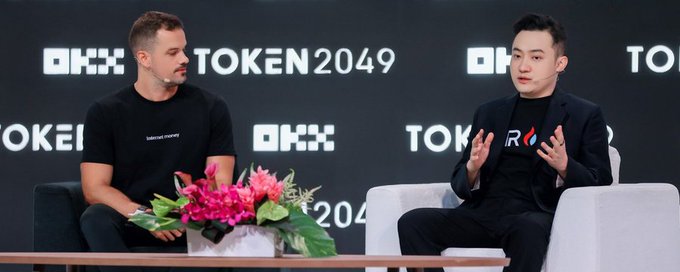 Two men sit on stage at the TOKEN2049 conference, with the man on the right wearing a TRX-branded blazer and gesturing as he speaks, while the man on the left listens attentively. The "OKX" and "TOKEN2049" branding is visible in the backdrop, consistent with the event where Justin Sun participated in a fireside chat hosted by Kevin Follonier.