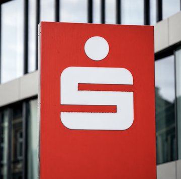 A red Sparkassen bank sign displaying the company's iconic "S" logo stands outside a building. The image visually ties to the news that Germany's largest banking group is expanding into Bitcoin and crypto trading services for its 40 million customers.