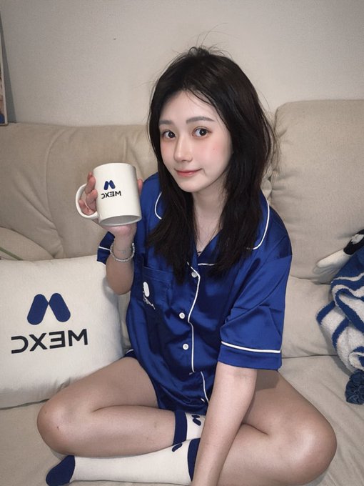 A young woman wearing blue MXC-branded pajamas sits on a couch holding a matching MXC mug, with an MXC pillow beside her.