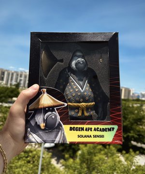 A hand holds up a "Degen Ape Academy Solana Sensei" NFT figurine in its packaging, depicting a wise ape in traditional samurai robes with a conical hat, connecting to the post's tribute to Degen Ape Academy as a foundational piece of Solana NFT culture.
