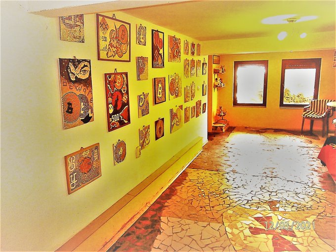 An indoor hallway features yellow walls covered in numerous small colorful paintings, a mosaic tile floor with a reflective puddle, and a timestamp of '12/03/2021' in the corner.