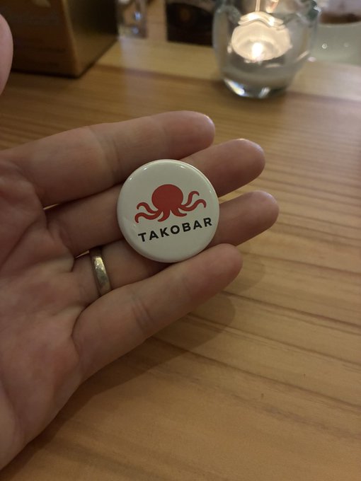 A hand holds a round white pin featuring a red octopus illustration with the text "TAKOBAR" beneath it, set against a wooden table with a lit candle in the background. The pin relates to the "たこバー" (Takobar) hashtag in the post, likely serving as a souvenir from the meetup.