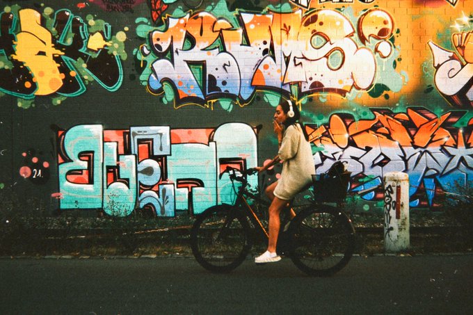 A person in a light dress and headphones rides a bicycle past a wall covered in vibrant, colorful graffiti art.