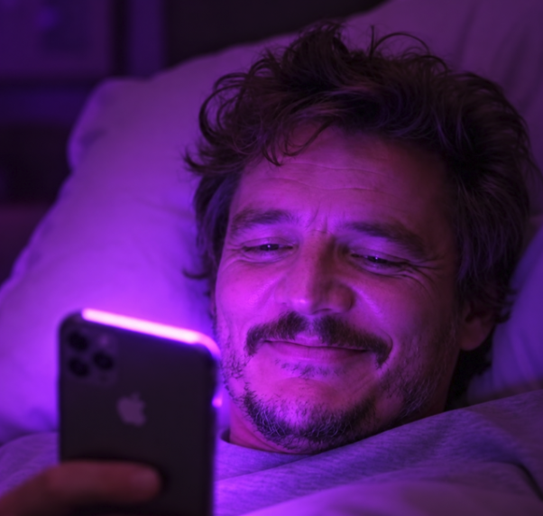 A man lying in bed smiling at an iPhone, with purple ambient lighting illuminating his face.