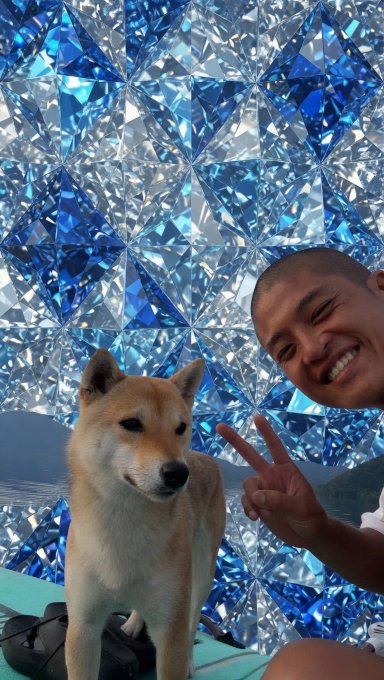 A smiling man poses with a peace sign next to a Shiba Inu dog in front of a sparkling blue crystal backdrop.