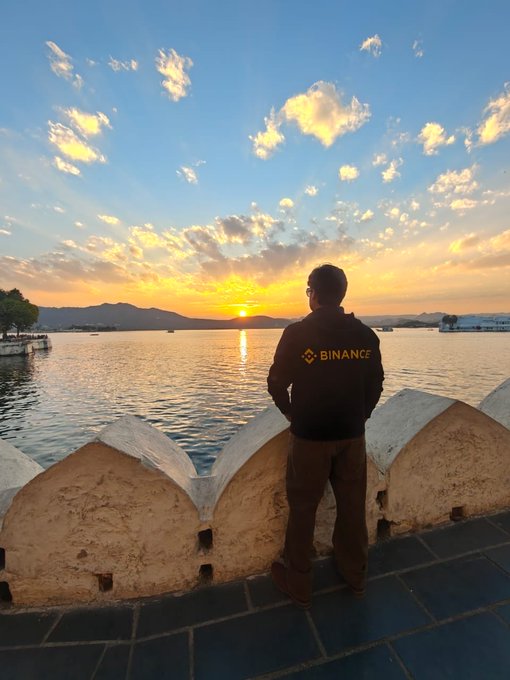 A person wearing a black Binance jacket stands on a stone pier at sunset, looking out over a lake with mountains in the background.