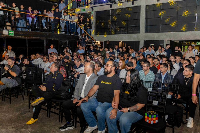 A large crowd of attendees fills a multi-level venue during Binance Day CDMX, with several participants wearing branded Binance apparel—including a man prominently sporting a "BINANCE" t-shirt—gathered for the crypto-focused event that drew over 400 Binancians for a full day of learning and networking.
