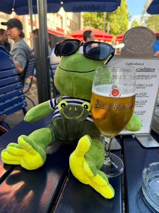 A green plush frog toy dressed in a striped shirt and sunglasses sits on an outdoor café table next to a glass of beer and a menu titled 'Gaffel-Kölsch Spezialitäten'.