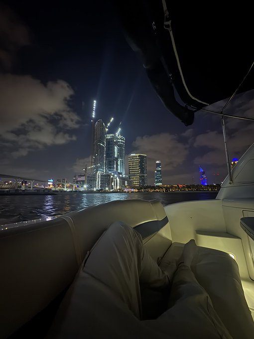 A person relaxes on a luxury yacht at night, legs stretched out as they gaze at the illuminated skyscrapers and city skyline across the water, capturing a moment of serene waterfront leisure.