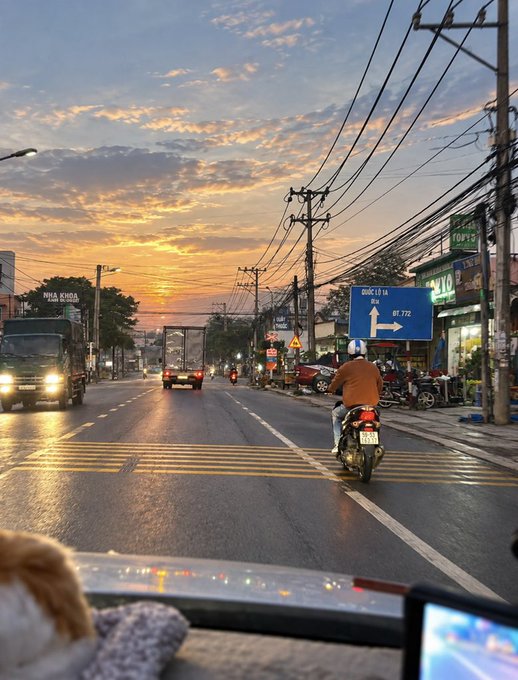 A view from inside a vehicle at sunset on a busy street, showing trucks and motorcycles with a blue sign indicating "QUỐC LỘ 1A" direction to "BỂN LỨC".