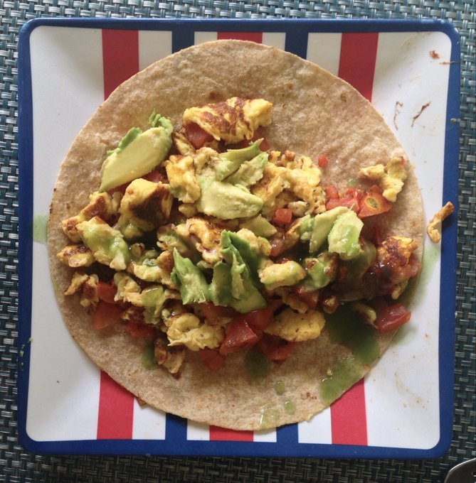 A breakfast tortilla on a red, white, and blue striped plate, filled with scrambled eggs, diced tomatoes, avocado chunks, and bacon, sitting on a woven blue placemat.