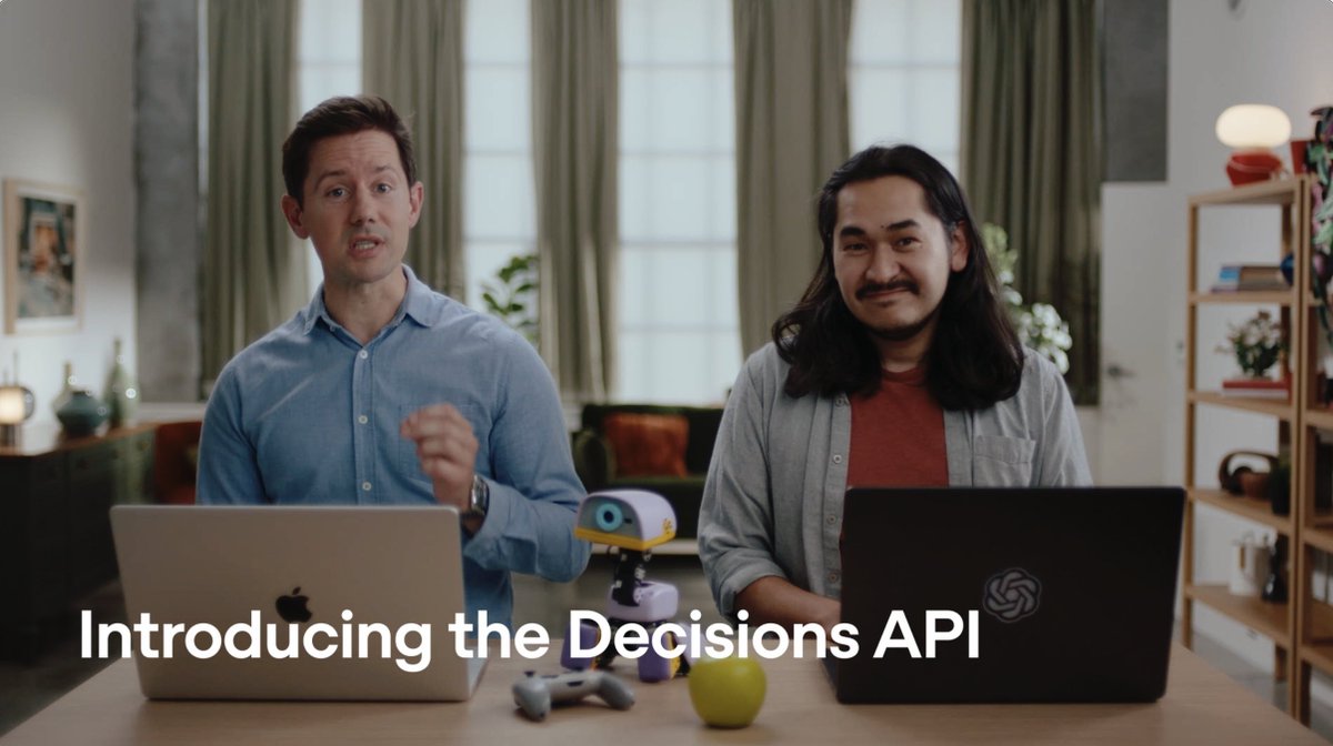 Two men sit at a desk with Apple and OpenAI laptops, a small robot, a game controller, and an apple, with the text 'Introducing the Decisions API' overlaid at the bottom.