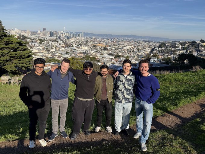 Six men pose together with their arms around each other on a grassy hill overlooking a city skyline, likely San Francisco.