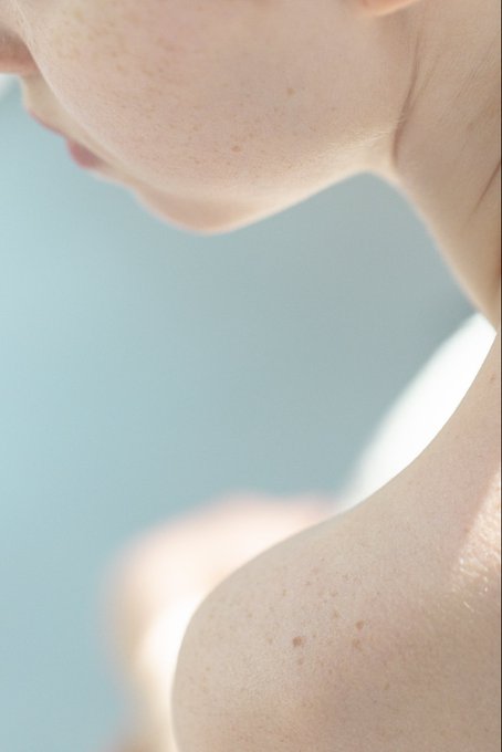 An extreme close-up photograph showing freckled skin and a sliver of red lips against a soft blue background.