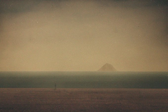 A muted, hazy landscape photograph featuring a flat field in the foreground, a calm sea or body of water in the middle, and a distant pyramid-shaped mountain under a pale, gradient sky.
