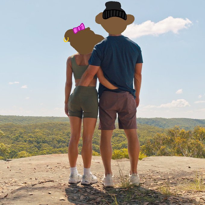 A couple stands together on a rocky overlook looking out at a forested landscape, with cartoon emoji masks covering their faces.