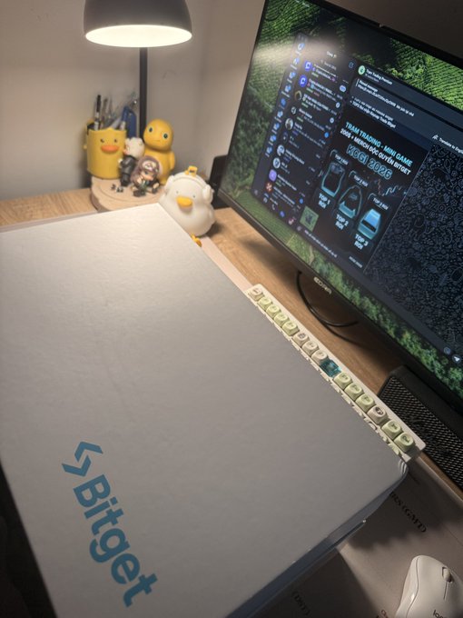 A desk setup featuring a closed silver laptop with 'Bitget' logo in blue, next to a monitor displaying Twitter, with yellow duck figurines and figurines in the background.