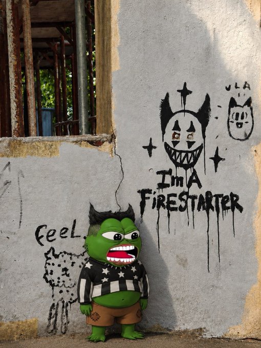 A green troll-style figurine with spiky black hair and checkered shirt stands against a weathered wall featuring graffiti that reads 'I'M A FIRESTARTER' with devil and cat drawings.