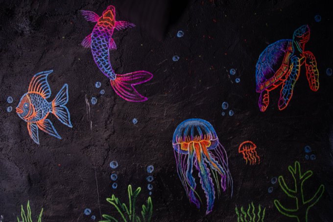 Neon-colored chalk drawings on a dark surface depicting a mermaid, fish, sea turtle, jellyfish, and seaweed in vibrant rainbow hues.