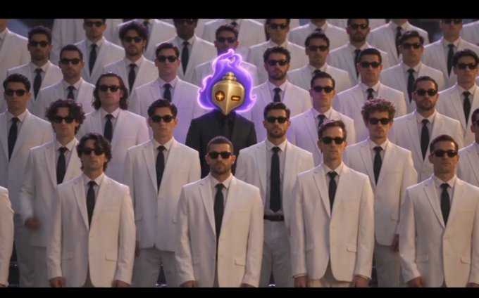 A crowd of men in matching white suits and black sunglasses surround a central figure wearing a glowing purple flame-headed mask, creating an eerie, uniform appearance reminiscent of a Secret Service-style scene.
