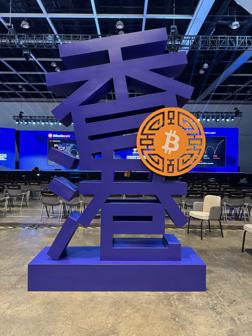 A large blue sculpture displays Chinese characters alongside an orange Bitcoin logo at what appears to be a Bitcoin conference venue.