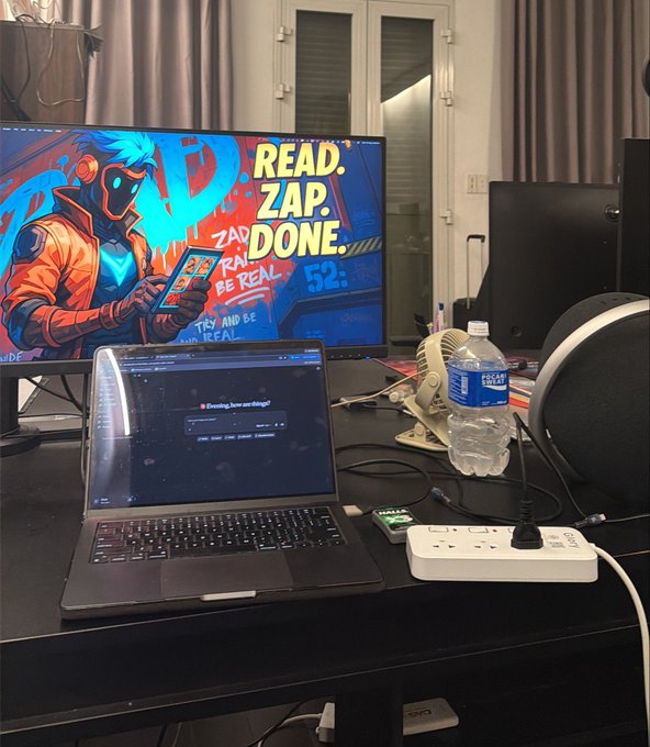 A desk setup featuring a monitor displaying 'READ. ZAP. DONE.' artwork of a futuristic character, a laptop with a video player, a water bottle, a power strip, and a fan on a dark desk.