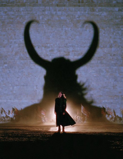 A lone figure in a long dark coat stands before a massive wall projection of a horned silhouette, with flags visible in the background.