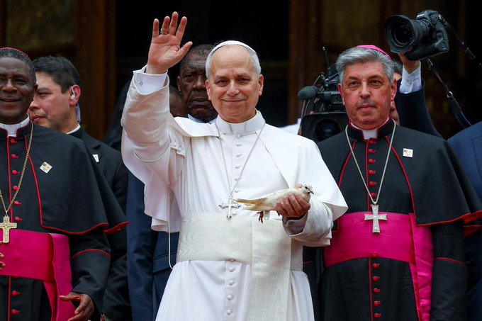 Pope Leo XIV (or a pope in white robes) waves while holding a white dove, flanked by clergy members in black with magenta trim.
