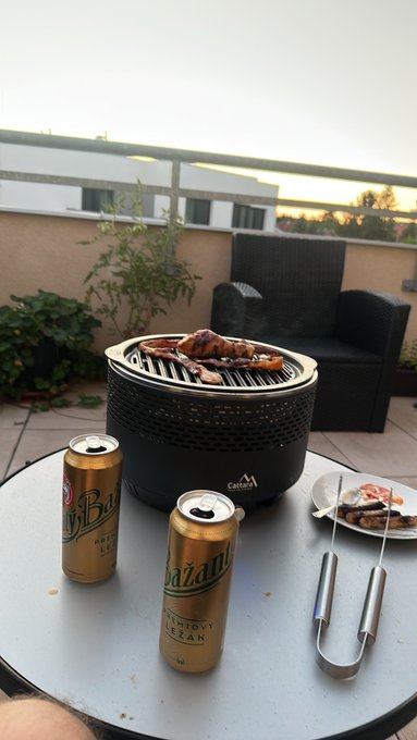 Two cans of Asahi beer on a table with a portable barbecue grill cooking meat, set on a balcony with wicker furniture during sunset.