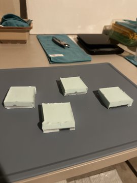 Four pale green, rectangular pixel-art objects arranged on a dark gray mat on a table, with cloth pouches and a pen visible in the background.