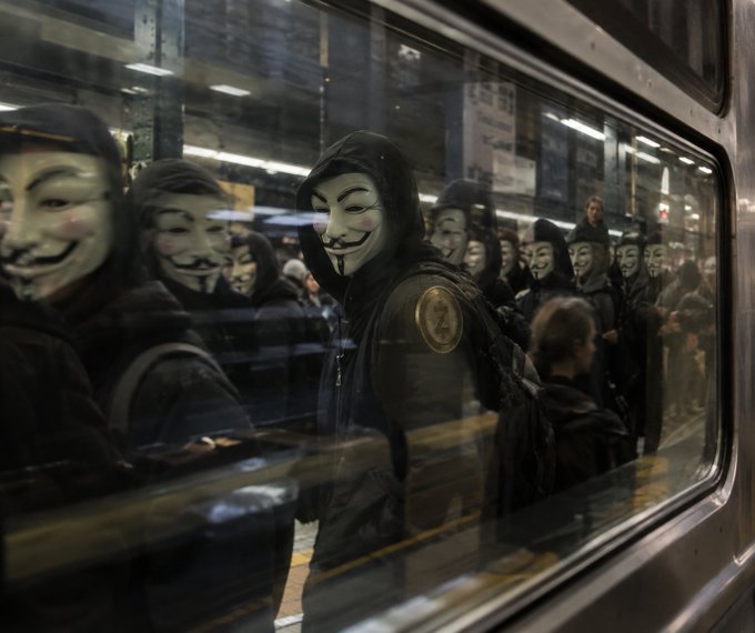Caption: A group of people wearing Guy Fawkes masks standing inside a subway car, seen through the window glass.