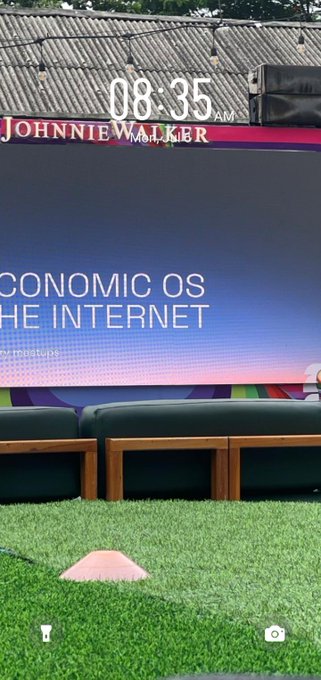 A phone screenshot at 08:35 AM showing a Johnnie Walker advertising sign that reads 'ECONOMIC OS' and 'THE INTERNET' with empty chairs in front.