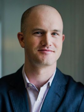 A portrait of a smiling, bald man wearing a dark blue suit jacket over a checkered shirt against a blurred background.