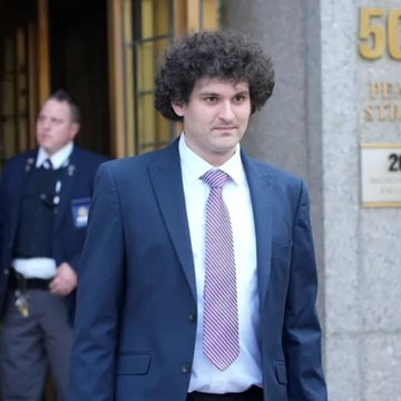 A man in a dark suit and pink tie exits a building near a "50" address marker, with a uniformed officer in the background. The image is associated with the denial of Sam Bankman-Fried's appeal by the Second Circuit Court of Appeals, affirming his fraud and conspiracy conviction.