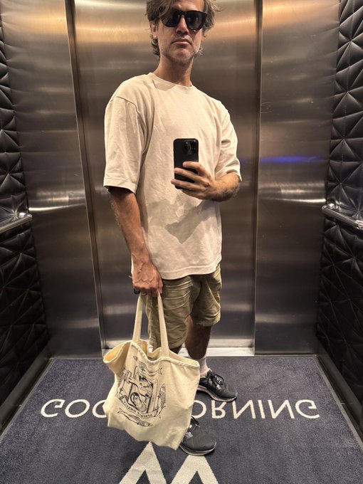 A man wearing sunglasses, a cream-colored t-shirt, and beige shorts stands in an elevator taking a mirror selfie, holding a canvas tote bag and a smartphone.