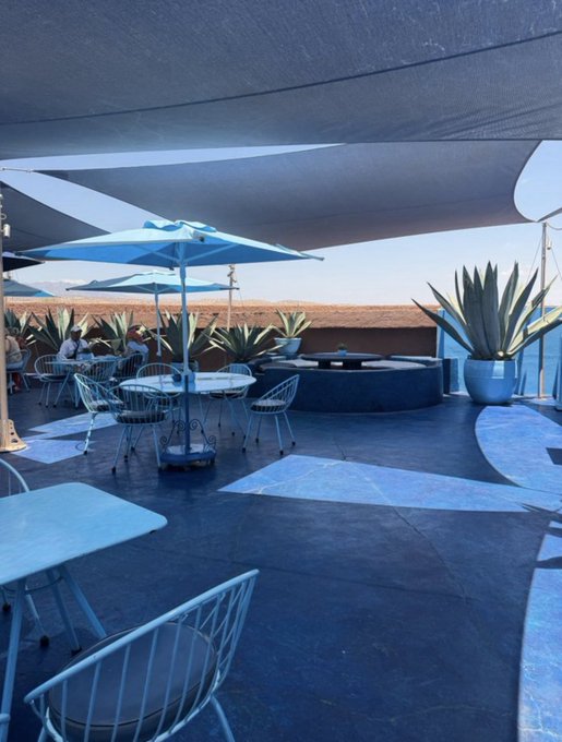 An outdoor patio dining area with blue umbrellas, wire chairs, and white tables set under large shade sails on a sunny day.