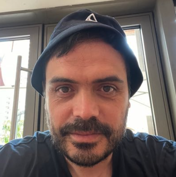 A close-up selfie of a bearded man wearing a dark blue bucket hat with a triangular logo.