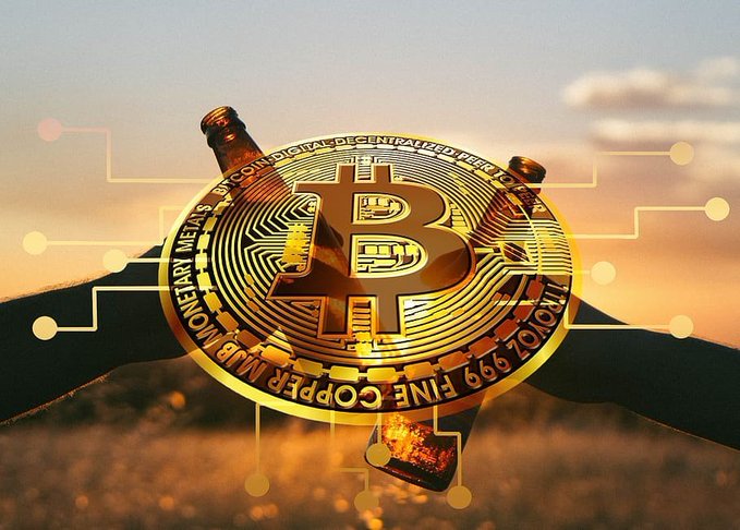 A golden physical Bitcoin coin is held aloft against a sunset sky, featuring the words 'BITCOIN DIGITAL DECENTRALIZED PEER TO PEER' and 'COPPER FINE .999' around its rim.