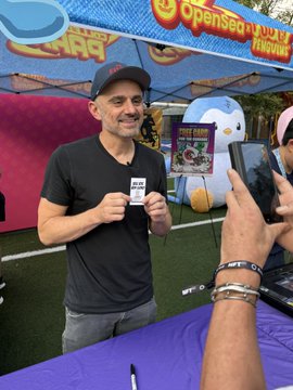 A man wearing a baseball cap and dark t-shirt stands at an outdoor event booth, holding up a card that appears to be from a cryptocurrency project, while another person photographs him with a phone.