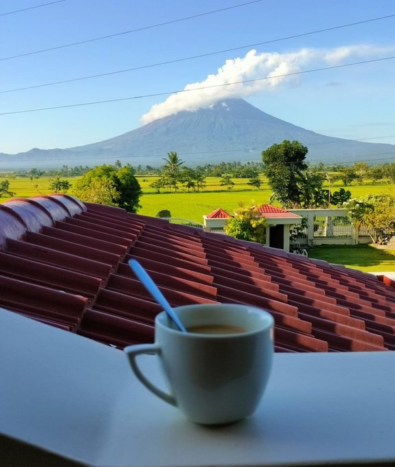 A scenic photo of a steaming white coffee mug in the foreground, overlooking a red-tiled roof with a volcano in the distance under a blue sky.