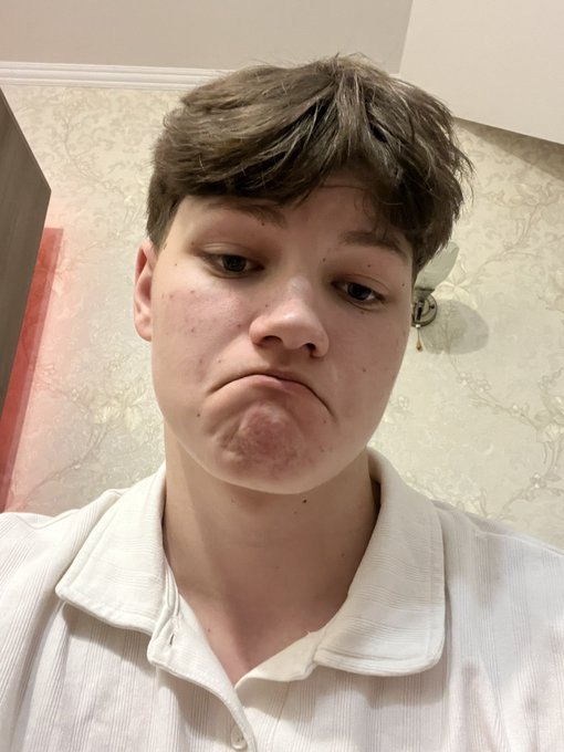 A young person with short brown hair making a frowning face at the camera, wearing a white collared shirt indoors.