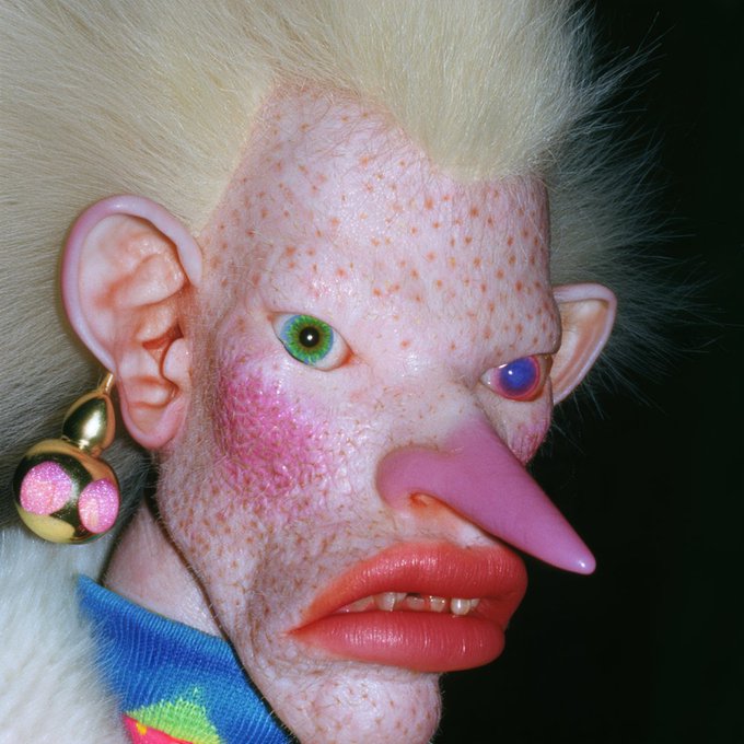 A close-up of a surreal, doll-like figure with a long pointed pink nose, heterochromatic eyes (green and red), freckled pale skin, wild white hair, and large pink earrings.