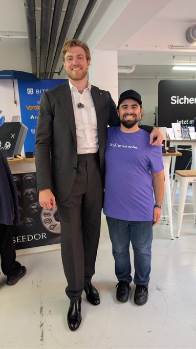 Two men stand together for a photo in an indoor event space; one wears a dark business suit and is quite tall, while the other wears a purple t-shirt with the text "to ❤ or not to be" and a black cap.