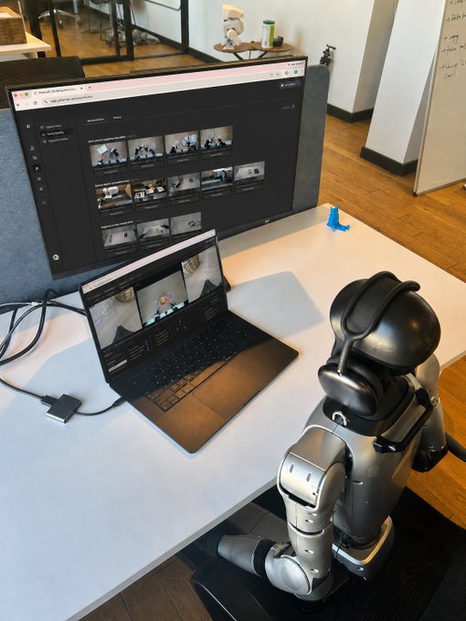 A humanoid robot sits at a desk in front of a laptop and monitor displaying the PrismaX AI platform interface, playfully positioned as if the robot is actively securing an early validator seat while the user merely scrolls. The setup visually reinforces the post's message about taking proactive action on app.prismax.ai rather than passively browsing.