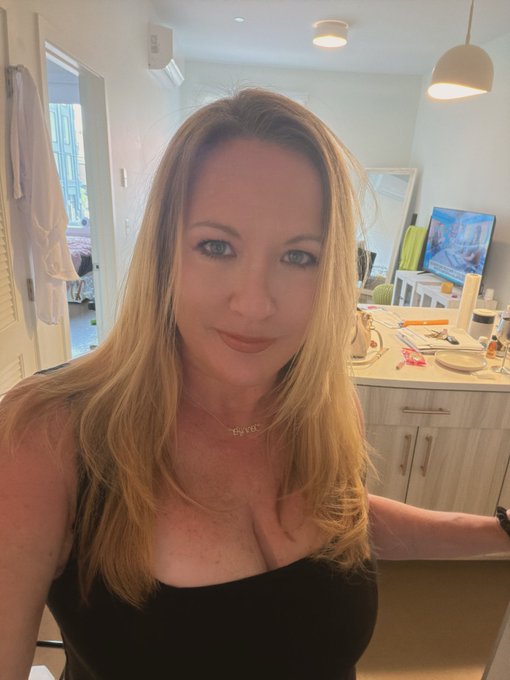 A selfie of a blonde woman wearing a black top and a gold 'love' name necklace, taken in what appears to be a bedroom with a vanity mirror and clothing visible in the background.