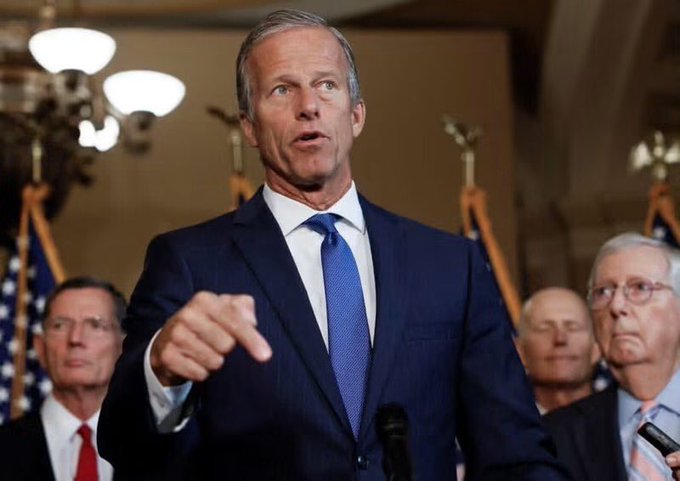 Senate Majority Leader John Thune, dressed in a dark blue suit and tie, speaks emphatically at a press conference, gesturing with his right hand while addressing reporters. The image illustrates the accompanying post about his plans to file cloture on the CLARITY Act before the August recess, with U.S. flags and other senators visible in the background of what appears to be a Capitol Hill setting.