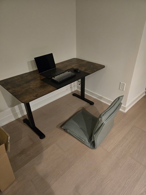 A minimalist home office setup featuring a dark wooden L-shaped desk with a laptop, keyboard, and mouse in the corner of a room with light-colored walls and flooring, accompanied by a cushioned floor seat.