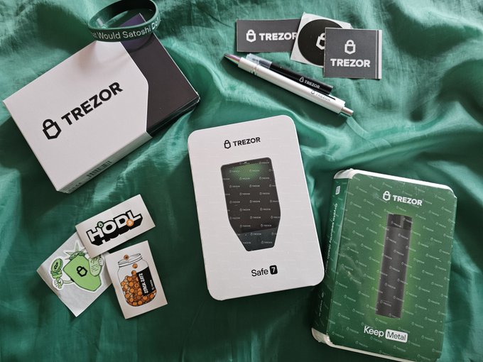 An unboxing scene with Trezor Safe 7 cryptocurrency hardware wallet packaging, stickers, pens, and a wristband laid out on a green satin fabric.