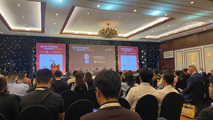 The image shows a large conference room filled with people sitting in chairs, watching a presentation being given by someone standing at a podium. The attendees are engaged and focused on the speaker's words. The room has a stage set up for presentations or discussions, indicating that this is an event where ideas and knowledge sharing take place.